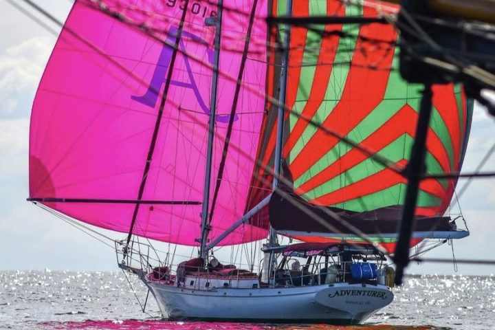 Sailboat with pink and green sails labeled 'ADVENTURER' on open water, viewed from behind.