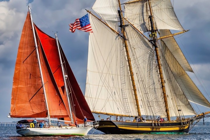 Two sailing ships on water, one with red sails and the other with cream sails, under a cloudy sky.