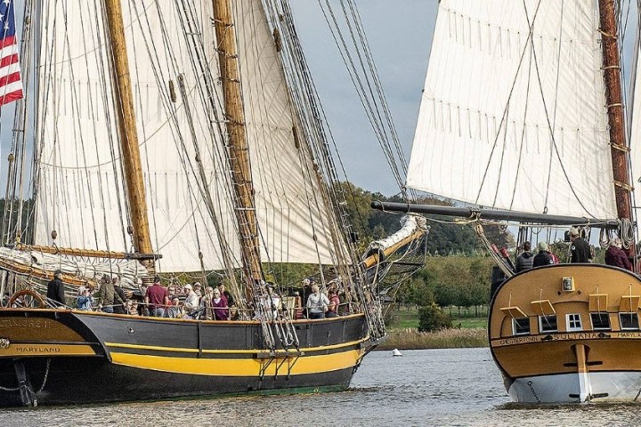 Two vintage sailing ships on water, one with an American flag, trees in background.