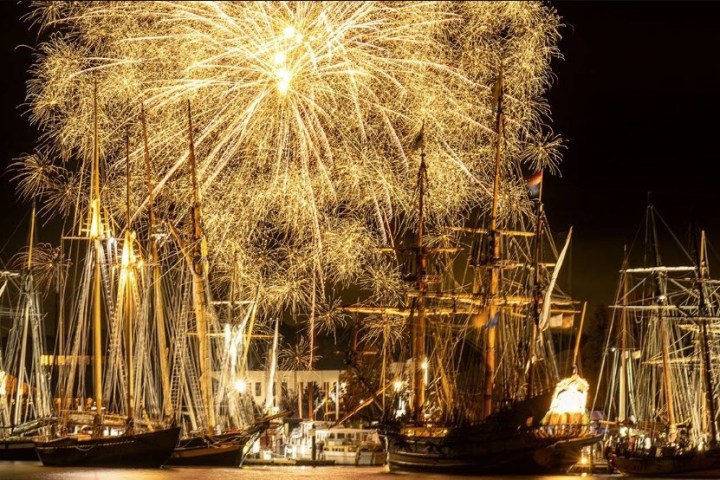 Fireworks illuminate tall ships at night in a harbor.