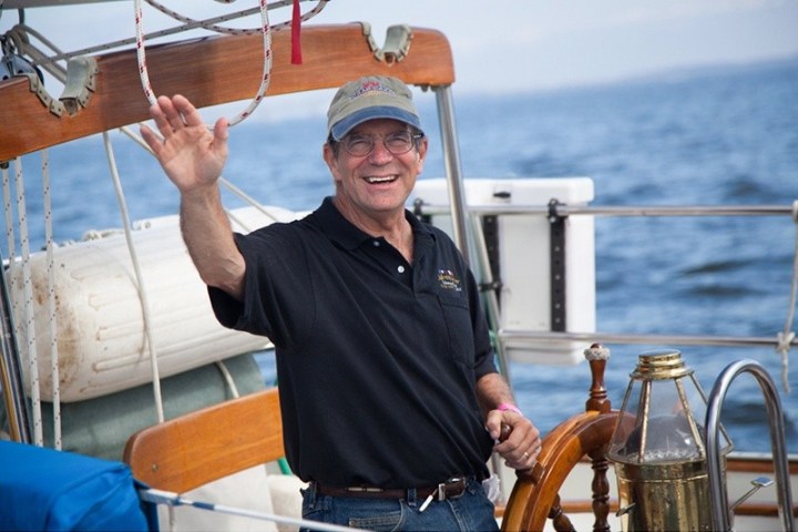 Man wearing a cap and glasses, smiling and waving on a boat.